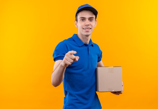 Young courier wearing blue uniform and blue cap happily pointing on and holds a box