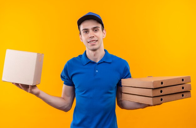 Young courier wearing blue uniform and blue cap holds a box on the left hand and three boxes on the right hand