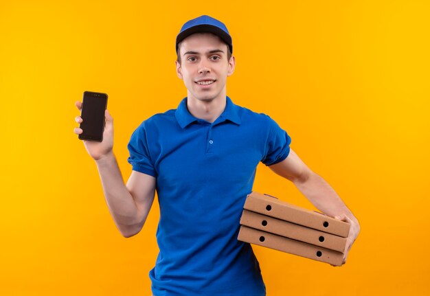 Young courier wearing blue uniform and blue cap holds boxes and a phone