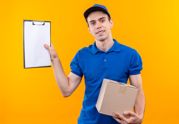 Young courier wearing blue uniform and blue cap holds a clipboard and box