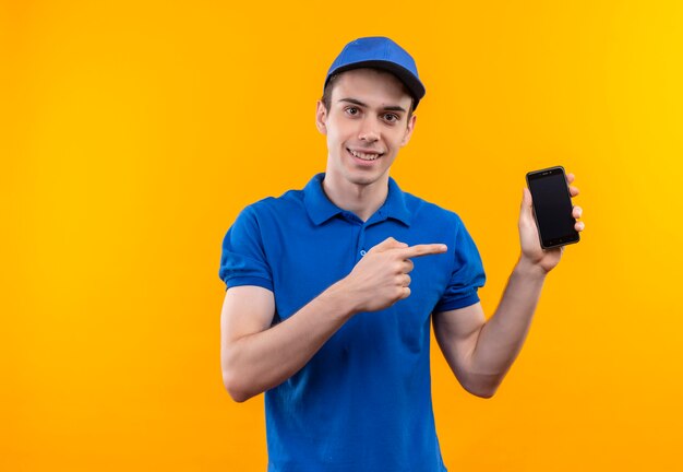 Young courier wearing blue uniform and blue cap points happy to he phone with forefinger