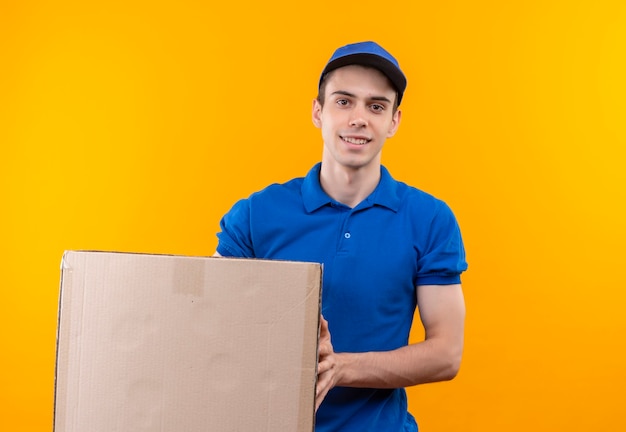 Young courier wearing blue uniform and blue cap smiles and holds a box