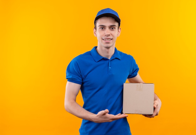 Young courier wearing blue uniform and blue cap smiles and holds a box