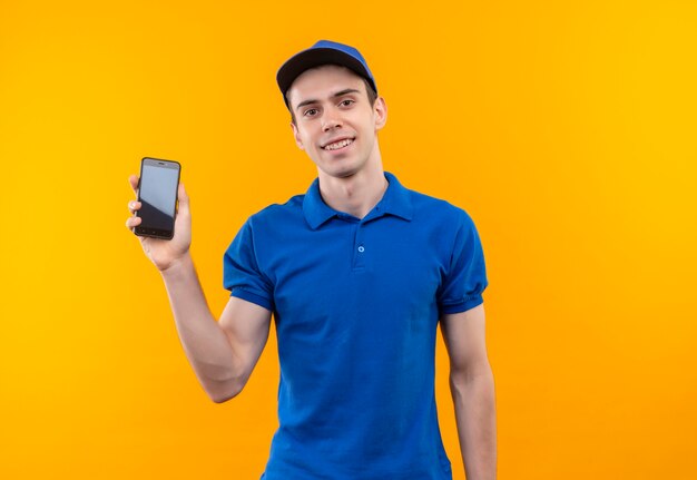 Young courier wearing blue uniform and blue cap smiling holds a phone