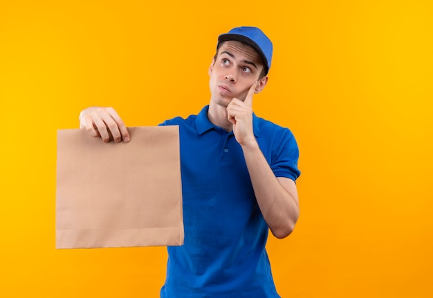 Young courier wearing blue uniform and blue cap thinking holds a bag