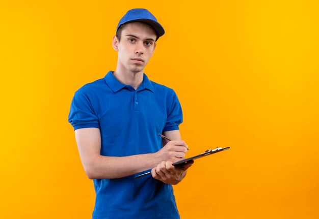 Young courier wearing blue uniform and blue cap writes on the clipboard