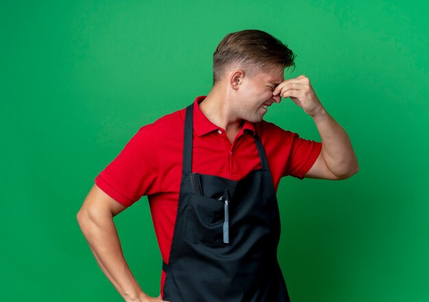 Young crying blonde male barber in uniform holds nose looking at side isolated on green space with copy space