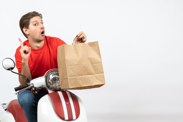 Young curious courier guy in red uniform sitting on scooter holding paper bag pointing up on white wall