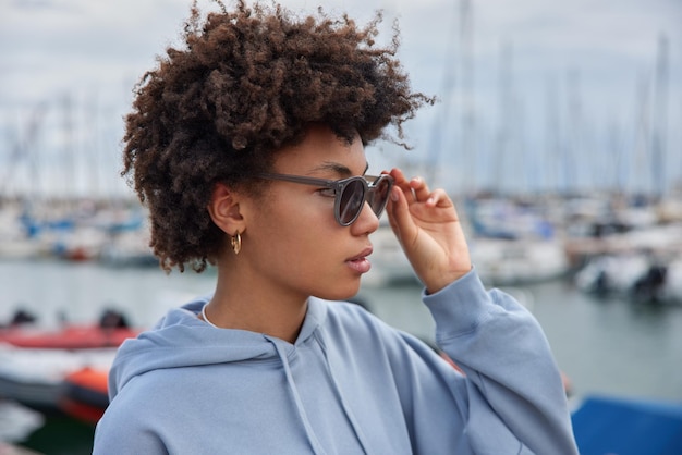 Free photo young curly female tourist wears sunglasses and sweatshirt focused into distance strolls at embankment going to have cruise on ship curly haired female stands outdoors in harbor. holiday concept