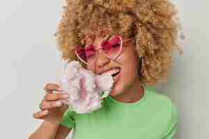 Free photo young curly haired pretty woman eats delicious cotton candy enjoys sweet snack wears pink heart sunglasses and casual green t shirt isolated over white background teenage girl bites candy floss