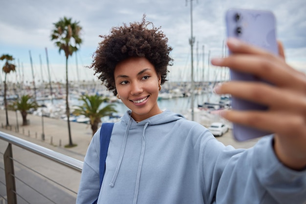 La giovane ragazza adolescente dai capelli ricci fa selfie sui sorrisi moderni dello smartphone indossa piacevolmente la felpa con cappuccio porta le pose dello zaino contro il porto si trova sul molo trascorre il tempo libero all'argine