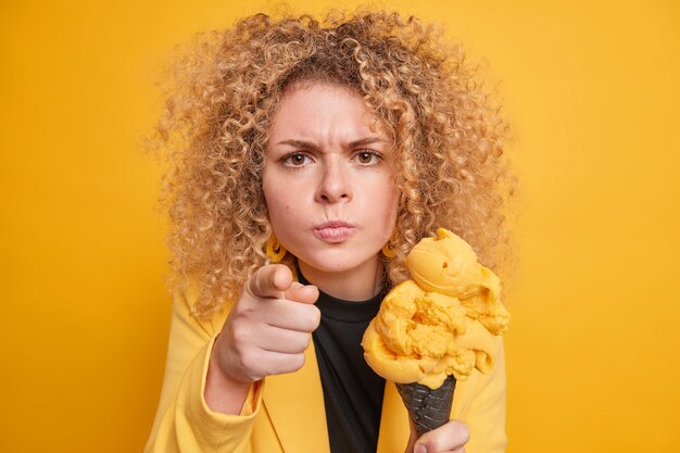 Young curly haired woman looks angrily and points directly  blames you holds delicious ice cream eats yummy summer dessert dressed in formal clothes isolated over yellow wall.