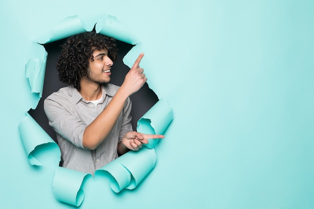 Young curly handsome man pointed on side from hole on green paper