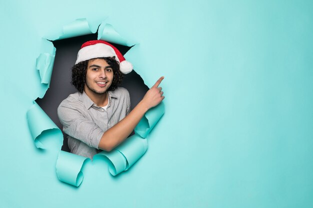 Young curly handsome man wear in santa hat pointed side from hole on green paper