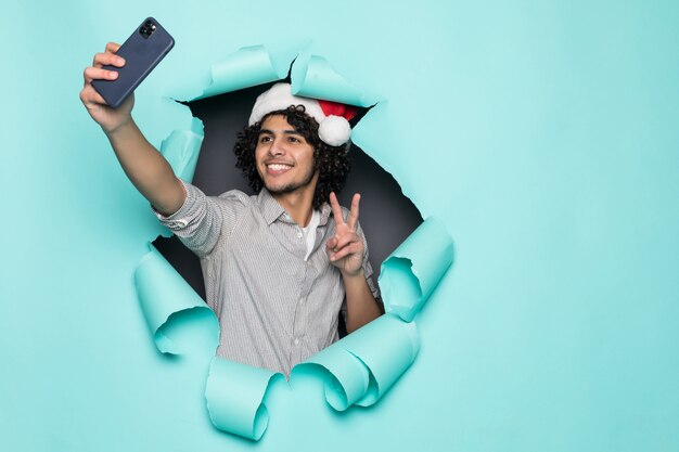 Young curly handsome man wear in santa hat take selfie from hole on green paper