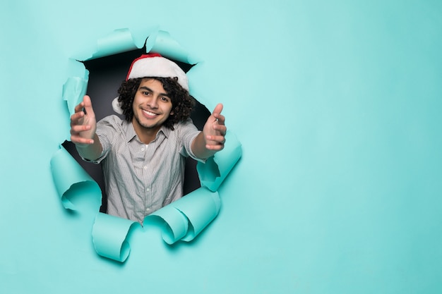 Young curly handsome man wear in santa hat with thumbs up from hole on green paper