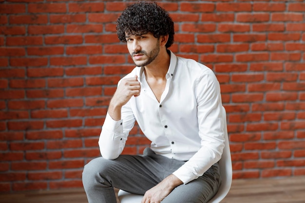 Young curly handsome in white shirt sitting on white chair. High quality photo