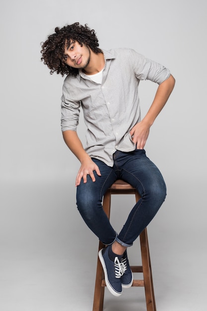 Young curly man sitting on a studio chair isolated on white wall