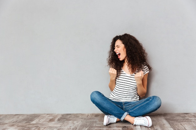 Free photo young curly woman in casual clothes expressing delight while sitting in lotus pose on the floor clenching her fists and shouting in joy over grey wall