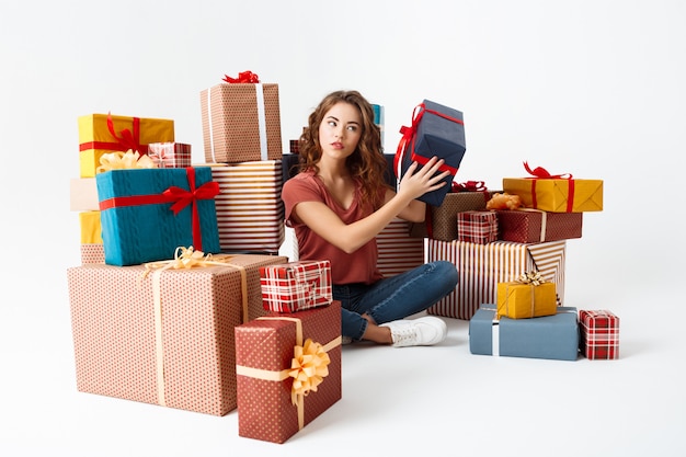 Young curly woman sitting on floor among gift boxes guessing what is inside