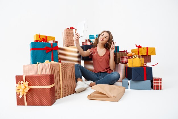Young curly woman sitting on floor among gift boxes making selfie with tablet