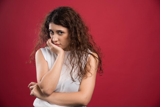 Young curly woman touching gently her face with raised hand on red. 