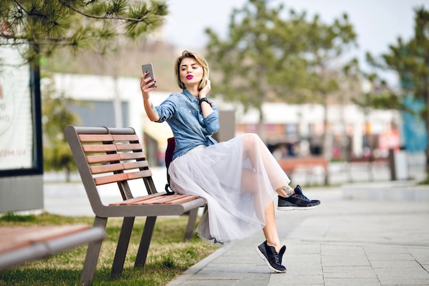 短い髪と唇にキスでselfieを作る木製のベンチに座っている明るいピンクの唇を持つ若いかわいい金髪の女性