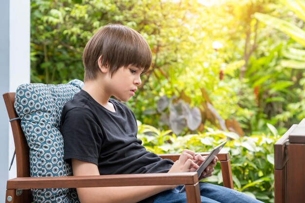 Young cute Caucasian boy using digital tablet sitting on couch in garden at home