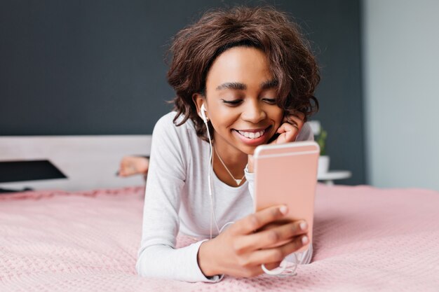 Young cute girl lying in bed, listening to music in earphones, looking at pink smartphone, texting with friends, web searching. Wearing nice gray t-shirt with long sleeves.