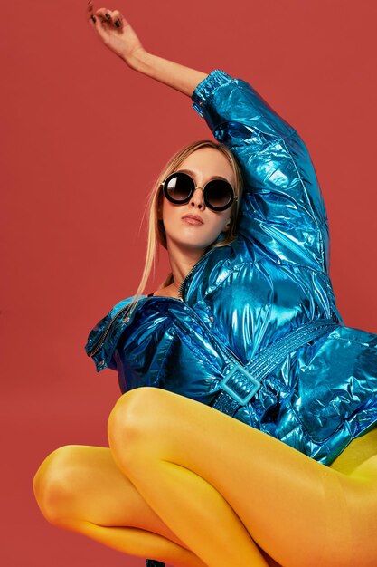 Young cute nice blonde girl in blue jacket sits in a fetal position in studio before camera, red background. Side view, concept art photo shoot