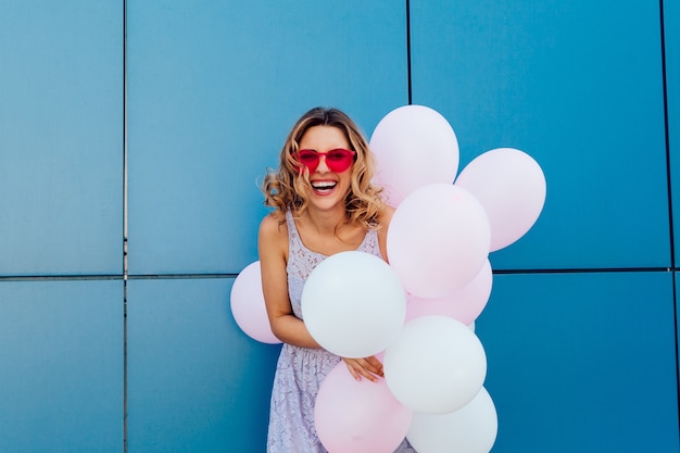 Young cute woman holding a bunch of air balloons, wearing pink sunglasses