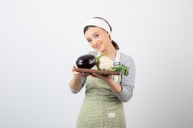 Молодая милая женщина-модель держит деревянную тарелку с баклажанами и цветной капустой