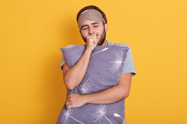 Young dark haired and bearded male yawning and covering mouth with his fist, keeps eyes closed, hugs a pillow