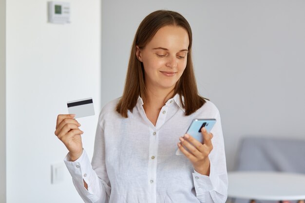 Young dark haired female wearing white shirt showing credit card and entering data in smart phone for online payments, looking at device screen with positive expression.