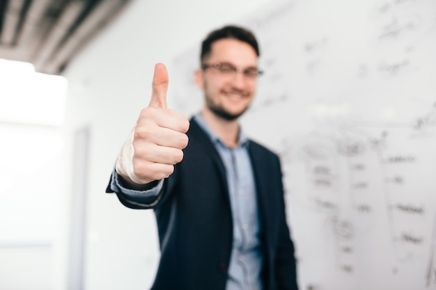 Free photo young dark-haired man in glasses is standing near whiteboard in office. he wears blue shirt and dark jacket. focus on front on his sign with hand.