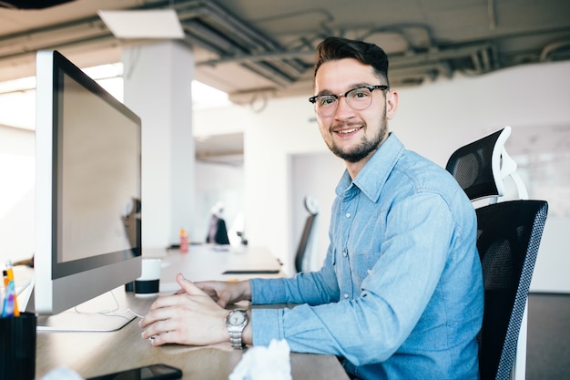 Молодой темноволосый мужчина в очках работает с компьютером на своем рабочем столе в офисе. Он носит синюю рубашку и улыбается в камеру. Вид сбоку.