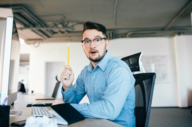 メガネの若い黒髪の男は、オフィスの職場に座っています。彼は青いシャツを着ています。彼は鉛筆を持っているし、カメラを見ています。