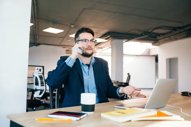 Young dark-haired man is working  at the table in office. He wears blue shirt with black jacket. He is speaking on phone and looks lost.