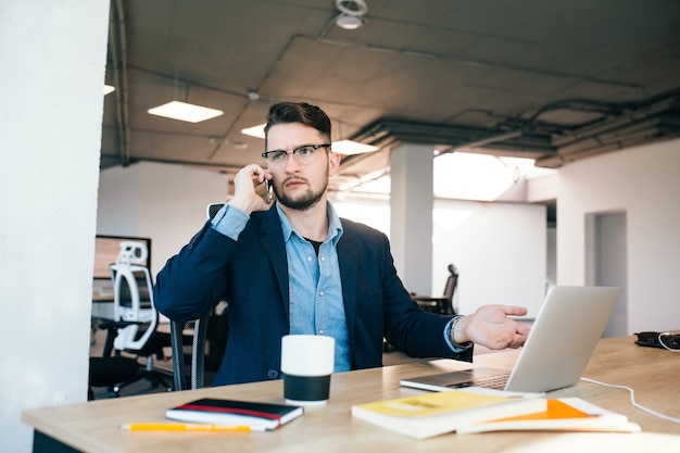 Young dark-haired man is working  at the table in office. He wears blue shirt with black jacket. He is speaking on phone and looks upset.