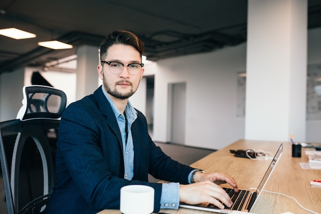 Young dark-haired man is working at the table in office. He wears blue shirt with black jacket. He is typing on laptop and looking to the camera.