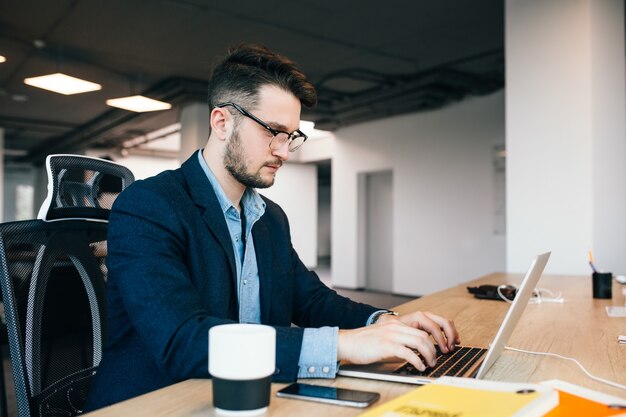 검은 머리 젊은이 사무실에있는 테이블에서 일하고있다. 그는 검은 색 재킷에 파란색 셔츠를 입는다. 그는 노트북에 타이핑하고 있으며 바쁘게 보입니다.