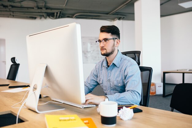 Young dark-haired man is working with  computer  at his desktop in office. He wears blue shirt and looks busy.