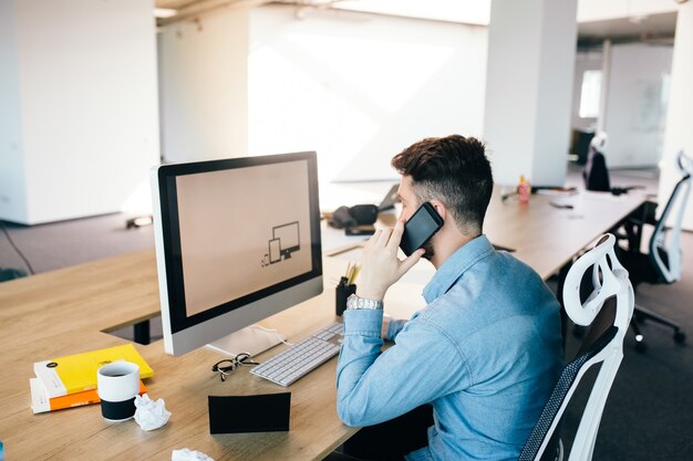 若い黒髪の男はコンピューターを使用して、オフィスの彼のデスクトップで電話で話しています。彼は青いシャツを着て、忙しく見えます。