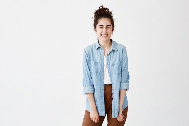 Young dark-haired woman clenches teeth in joy, dressed in denim shirt over white top, having good mood. Face expression, feelings and emotions