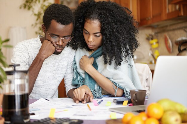 Young dark-skinned couple managing finances, sitting at kitchen table with stressed looks