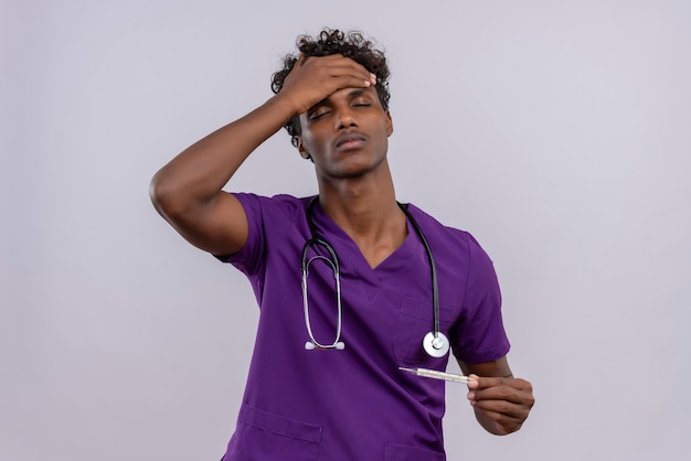 A young dark-skinned doctor with curly hair wearing violet uniform with stethoscope holding thermometer while keeping palm on forehead 