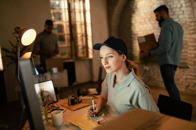 Young deliverer filling data on package label while using computer in the office