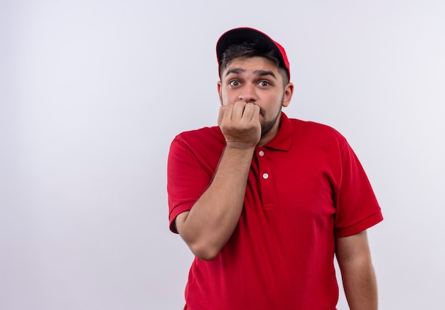 Young delivery boy in red uniform and cap stressed and nervous biting nails 