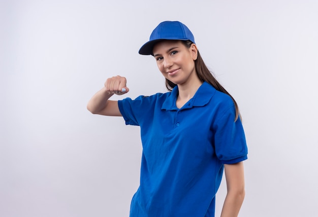 Young delivery girl in blue uniform and cap clenching fist making greeting gesture smiling friendly 