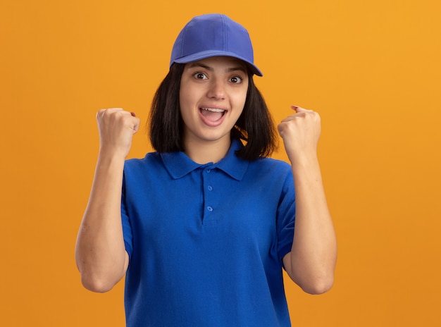 Free photo young delivery girl in blue uniform and cap clenching fists hapy and excited standing over orange wall